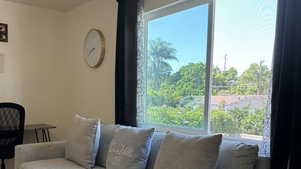 A living room with a grey sofa, a clock on the wall, and a view of greenery outside. - CocoVillaSB Apartment #4 (Santa Barbara)