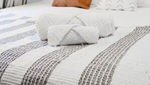Una cama bien tendida con un edredón blanco y una manta estampada, un almohadón marrón y un almohadón blanco con diseño de estampilla.