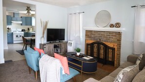 Una sala de estar con un sofá, una mesa de centro y una chimenea. Se ve una televisión y una cocina al fondo.