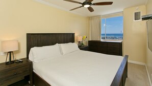 A hotel room with a large bed, a ceiling fan, a nightstand with a lamp and remote, a dresser, and a window with a view of the city. - Renovated Oceanfront Condo (Honolulu)