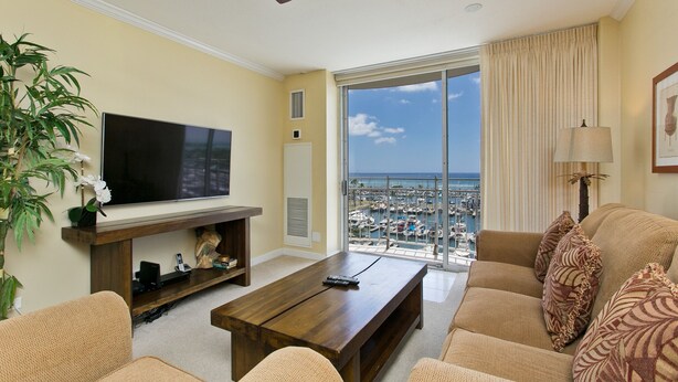 A living room with a TV, a coffee table, and a sofa. There is a view of a marina through large windows. - Renovated Oceanfront Condo (Honolulu)