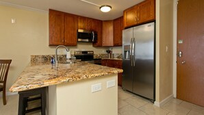 A kitchen with wooden cabinets, a stainless steel refrigerator, a granite countertop, and a built-in microwave. - Renovated Oceanfront Condo (Honolulu)