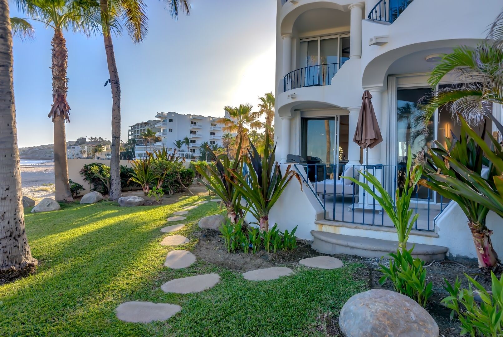 Um hotel moderno com varanda, um jardim bem cuidado com palmeiras e pedras, e vista para a praia e outros edifícios ao fundo.