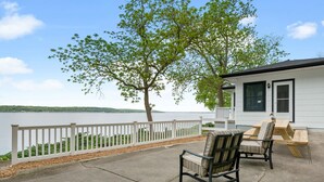 A patio with two chairs and a table, overlooking a lake with a white fence and a tree.