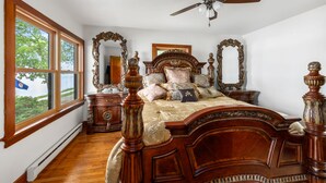 A bedroom with a large bed, ornate wooden headboard, two nightstands, a ceiling fan, and a window with a view of greenery.