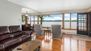 A spacious living room with a leather sofa, two armchairs, and a coffee table. A large window offers a view of a lake and distant hills.