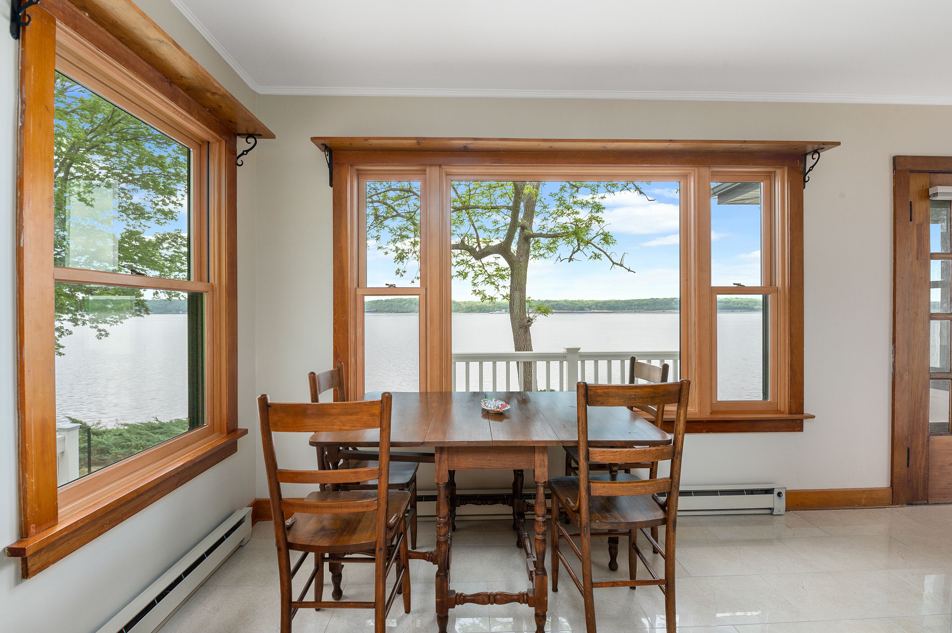 A dining area with a wooden table and chairs, large windows with a view of a lake and trees, and a white railing.