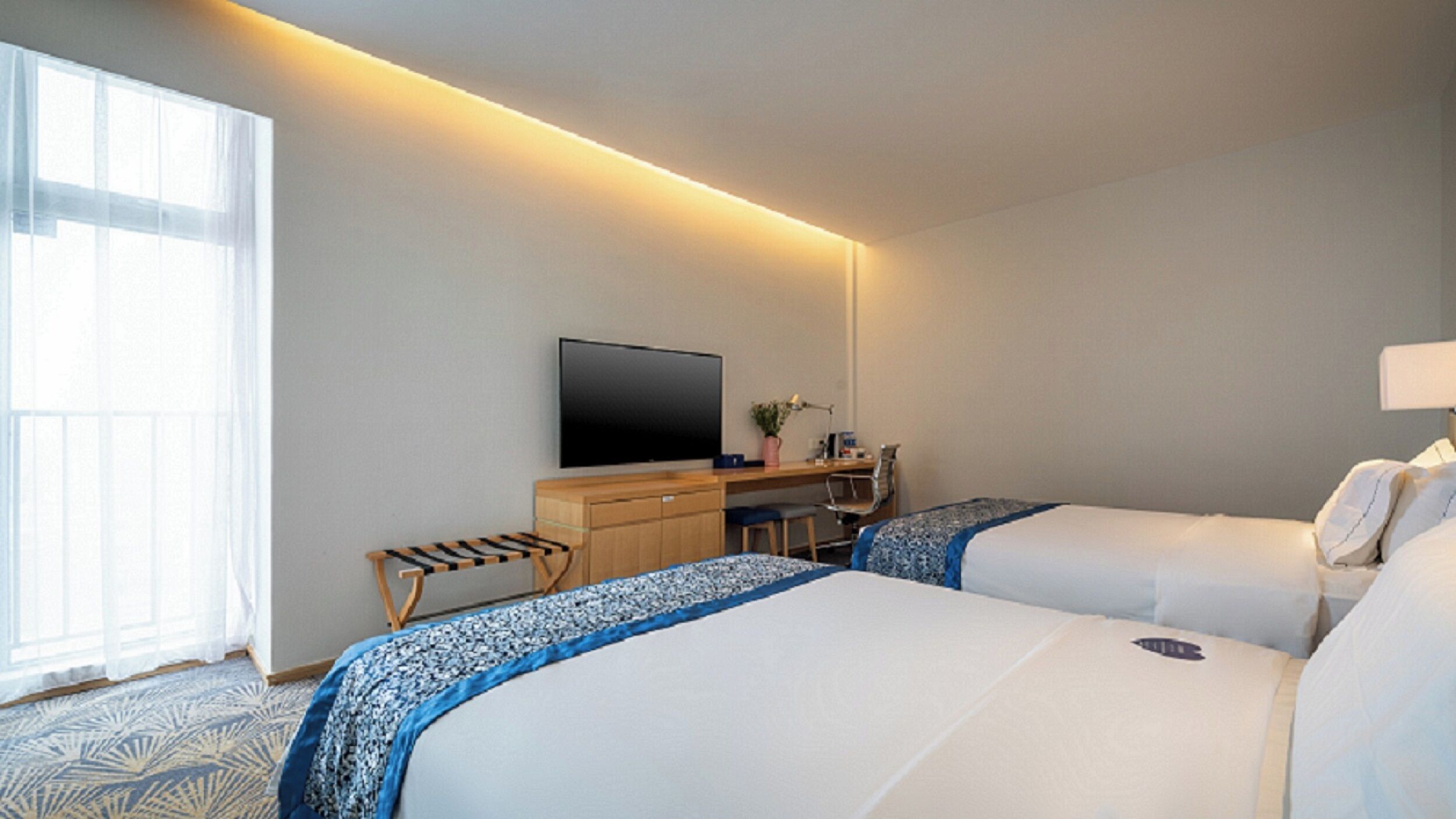 Ein Hotelzimmer mit zwei Betten, einem Schreibtisch, einem Fernseher und einem Fenster mit durchsichtigen Vorhängen.
