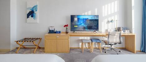 A hotel room with two beds, a desk with a computer, a chair, a TV displaying a welcome message, and a small table with a vase of flowers.
