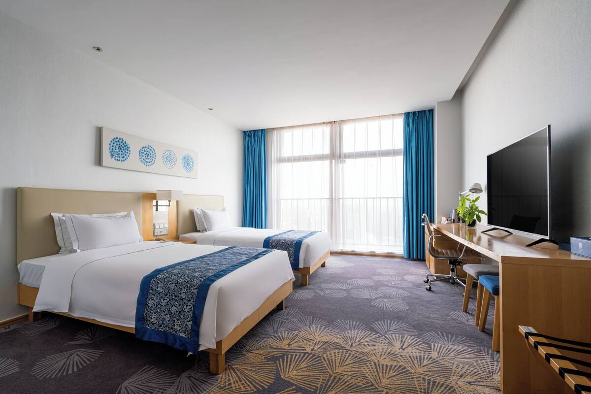 Une chambre d’hôtel avec deux lits, un bureau avec une chaise, une télévision et une grande fenêtre avec des rideaux.