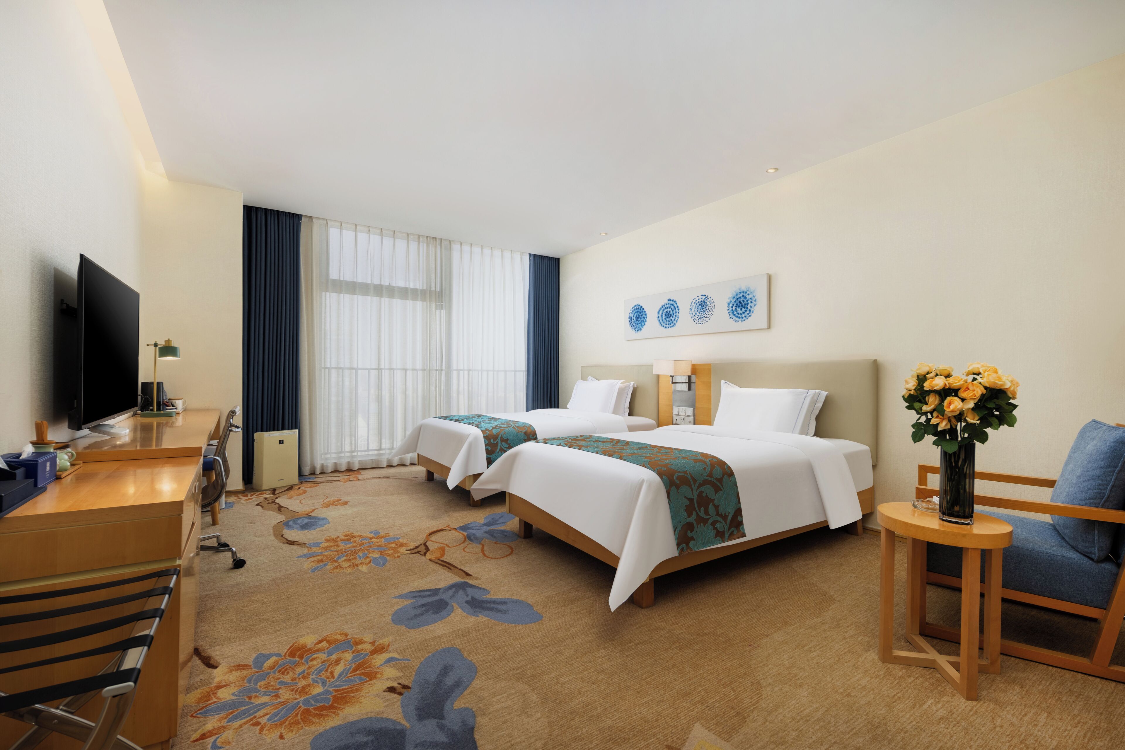 A hotel room with two beds, a desk with a TV, a chair, a small table with a vase of flowers, and a large window with curtains.