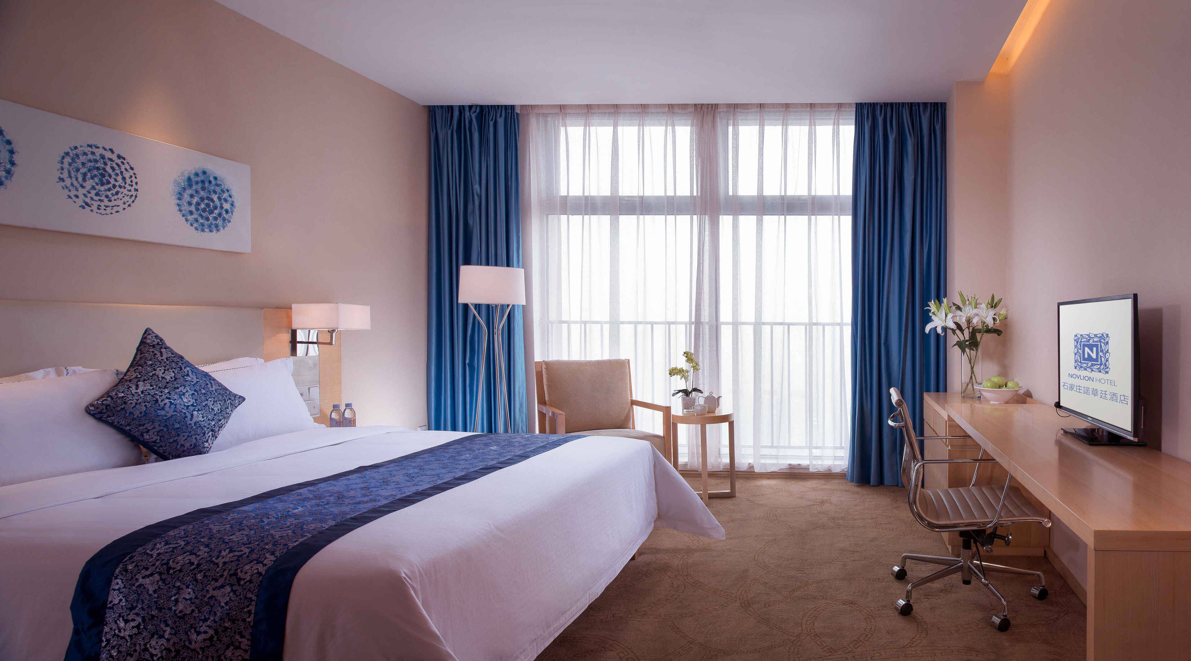 A hotel room with a large bed, a desk with a computer, a chair, a TV, and a window with blue curtains.
