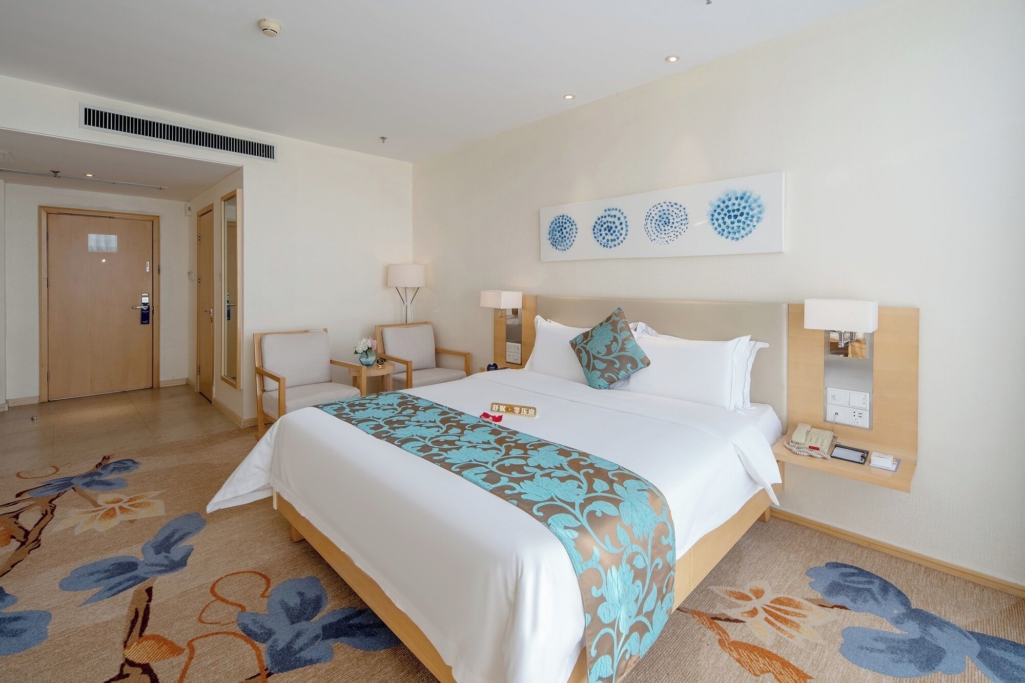 A hotel room with a bed, two chairs, a desk, a lamp, a phone, and a wall art piece.