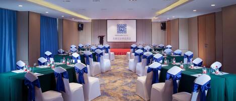 Une salle de conférence avec des rangées de chaises blanches ornées de rubans bleus, des tables avec des bouteilles d’eau et un écran affichant le logo de l’hôtel Novion.