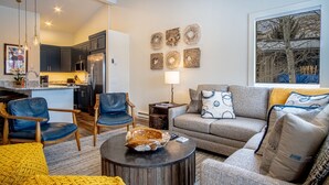 A modern living room with a grey sofa, blue and yellow armchairs, a coffee table, and a kitchen in the background. - Steps to Free Shuttle • 3-Min Ride to Ski Lifts & Ketchum (Ketchum)