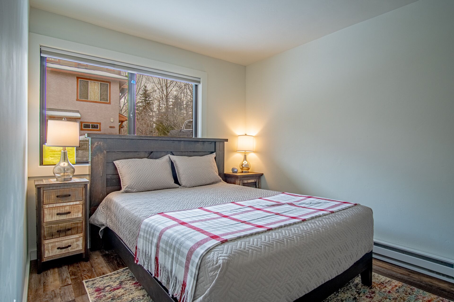 Een net opgemaakt bed met een geruit plaid, twee bedlampen, een nachtkastje met lamp en een raam met blinds.
