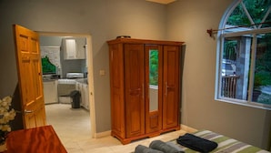 A bedroom with a bed, a wooden wardrobe, a window with a view of greenery, and an open door leading to a kitchen. - Majestic Villa, breathtaking view of famous Pitons, Ocean and Soufriere Town (Soufriere)