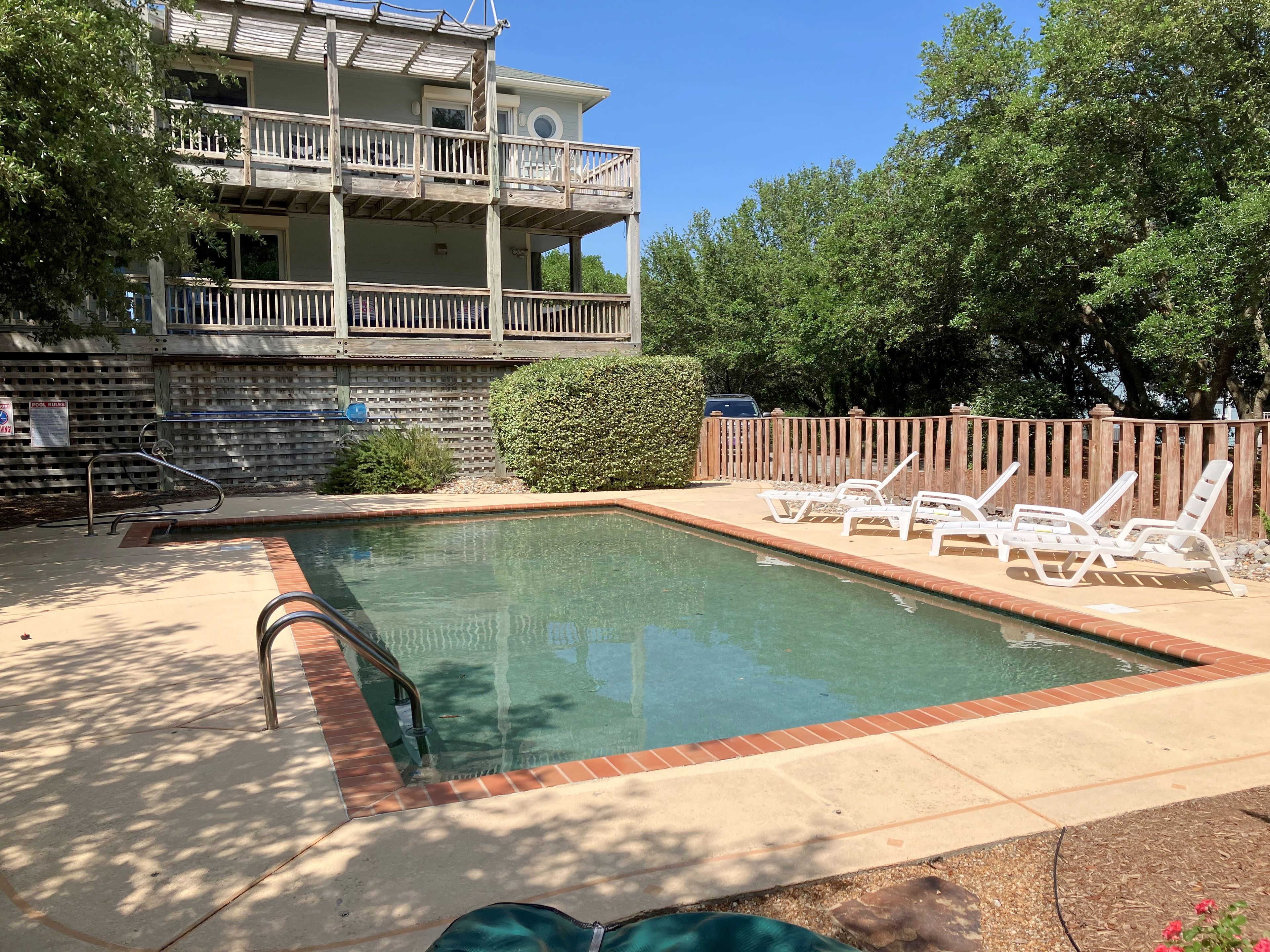 A swimming pool with a wooden deck, lounge chairs, and a multi-story building in the background.