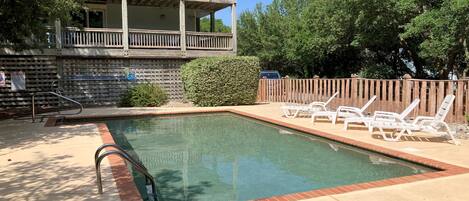 A swimming pool with a wooden deck, lounge chairs, and a multi-story building in the background.