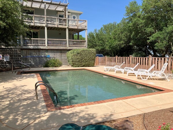 A swimming pool with a wooden deck, lounge chairs, and a multi-story building in the background.