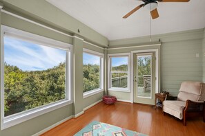 Ein Zimmer mit großen Fenstern, einem Deckenventilator, Holzboden und einem Korbstuhl.