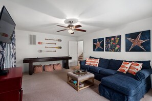 A living room with a dark blue sofa, a wooden coffee table, a ceiling fan, and wall art including starfish and jellyfish paintings. - Chase-n-Grace: Private Pool, Spacious Yard, Short Walk to Beach (Southern Shores)