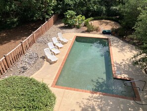 A rectangular pool with a wooden deck and lounge chairs, surrounded by a wooden fence and greenery.