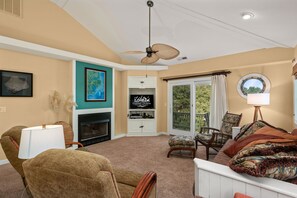 A living room with a fireplace, a ceiling fan, a television, and a view of the outdoors.
