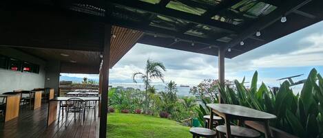An outdoor dining area with wooden tables and chairs, a grassy lawn, and a view of the sea.