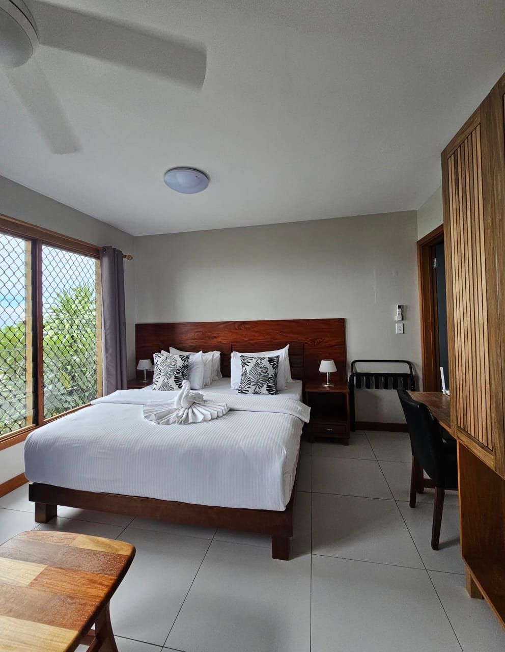 A hotel room with a large bed, a wooden headboard, a small table, a chair, and a view through a window.