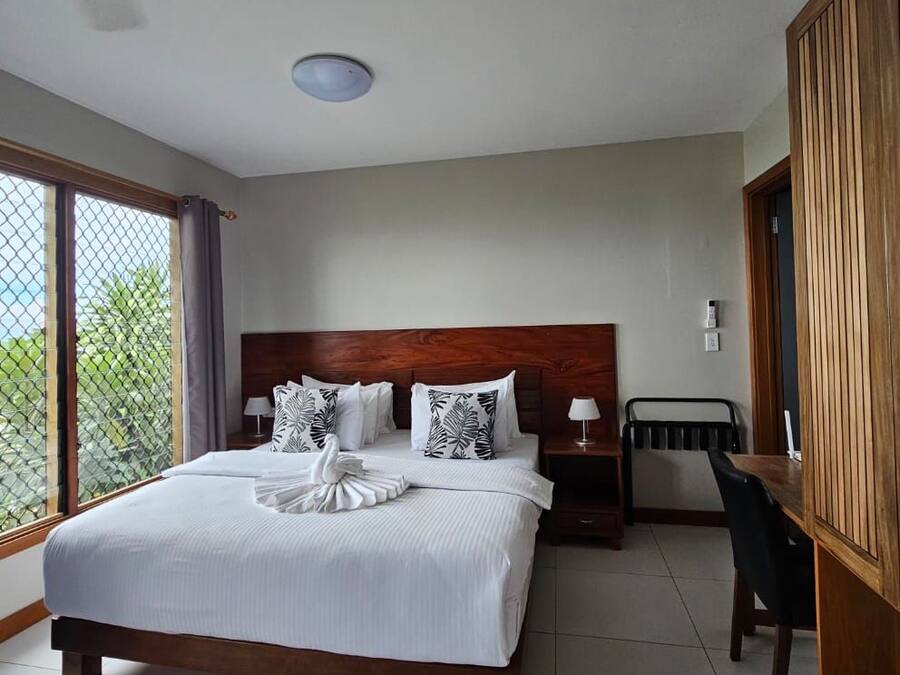 A hotel room with a large bed, a wooden headboard, a small table, a chair, and a view through a window.