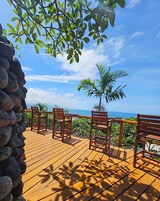 Um deck de madeira com cadeiras e uma mesa, com vista para a paisagem cênica do oceano e das montanhas.