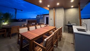 Ein Essbereich im Freien mit einem Holztisch und -stühlen, einem Grill und Blick auf die Skyline der Stadt in der Dämmerung.