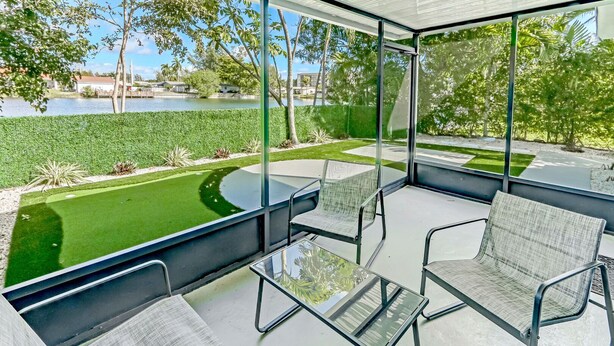 A modern outdoor seating area with glass tables and chairs, overlooking a well-maintained garden and a body of water. - Hallandale Collection: Renovated Professional Design & Lake View (Hallandale)