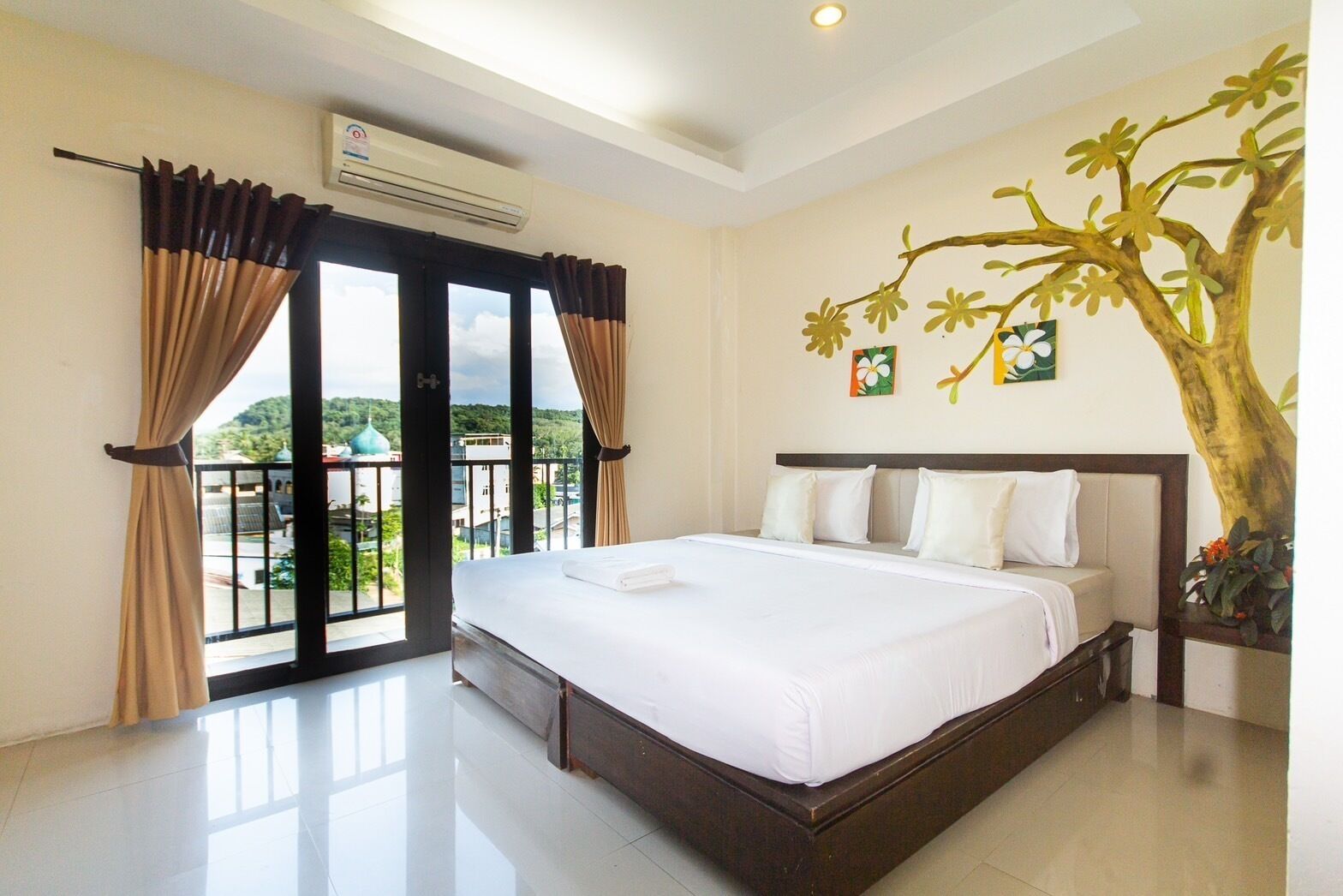A hotel room with a large bed, a balcony with curtains, an air conditioning unit, and a decorative tree painting.
