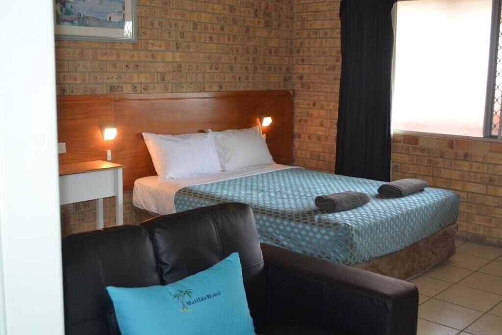 A hotel room with a brick-patterned wall, a wooden headboard, a bed with a blue patterned bedspread, two pillows, a nightstand with a lamp, and a chair with a pillow featuring a palm tree.