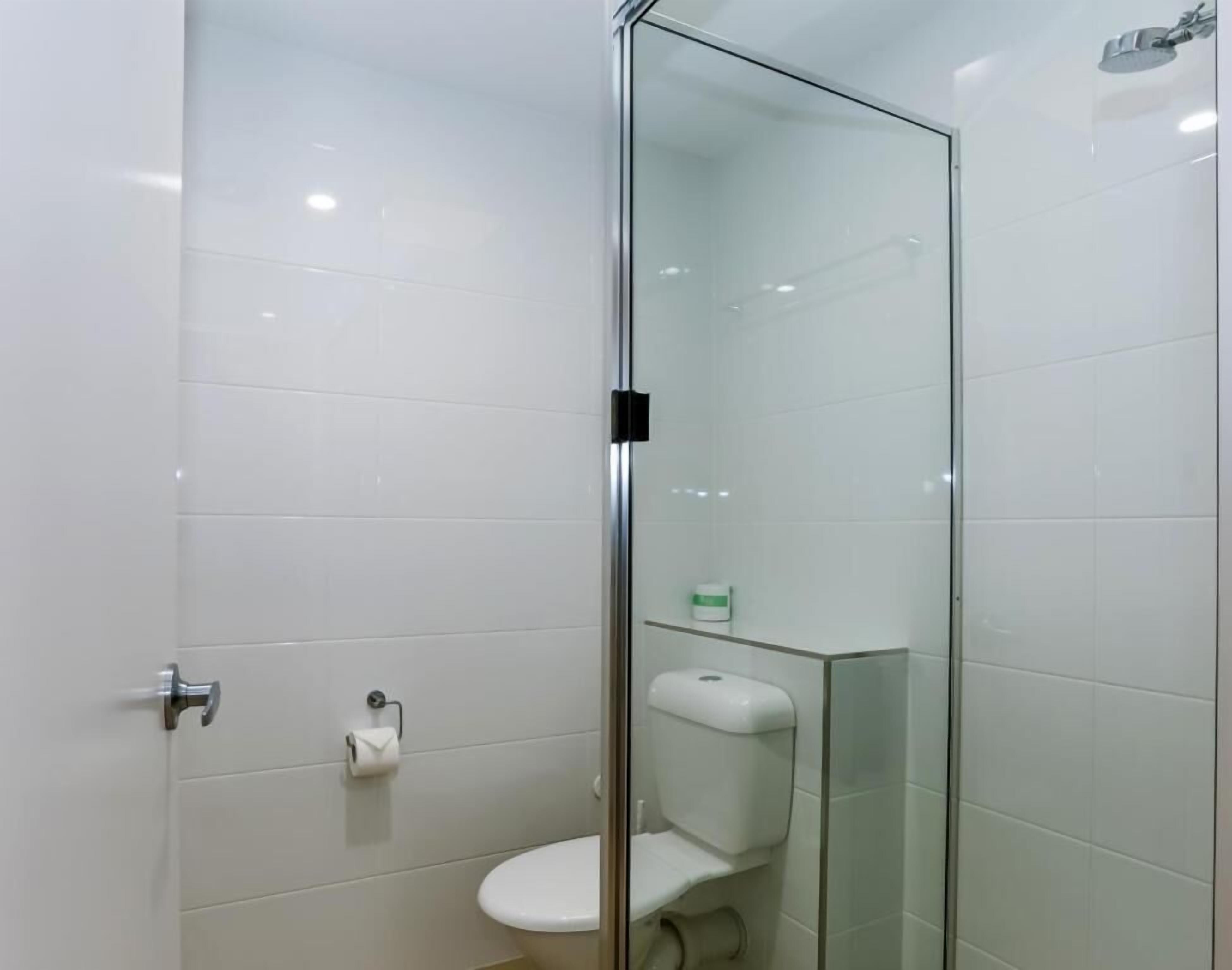 A modern bathroom with a glass shower enclosure, white tiles, a toilet, and a small shelf with toiletries.
