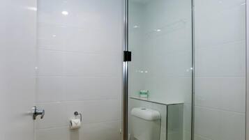 A modern bathroom with a glass shower enclosure, white tiles, a toilet, and a small shelf with toiletries.