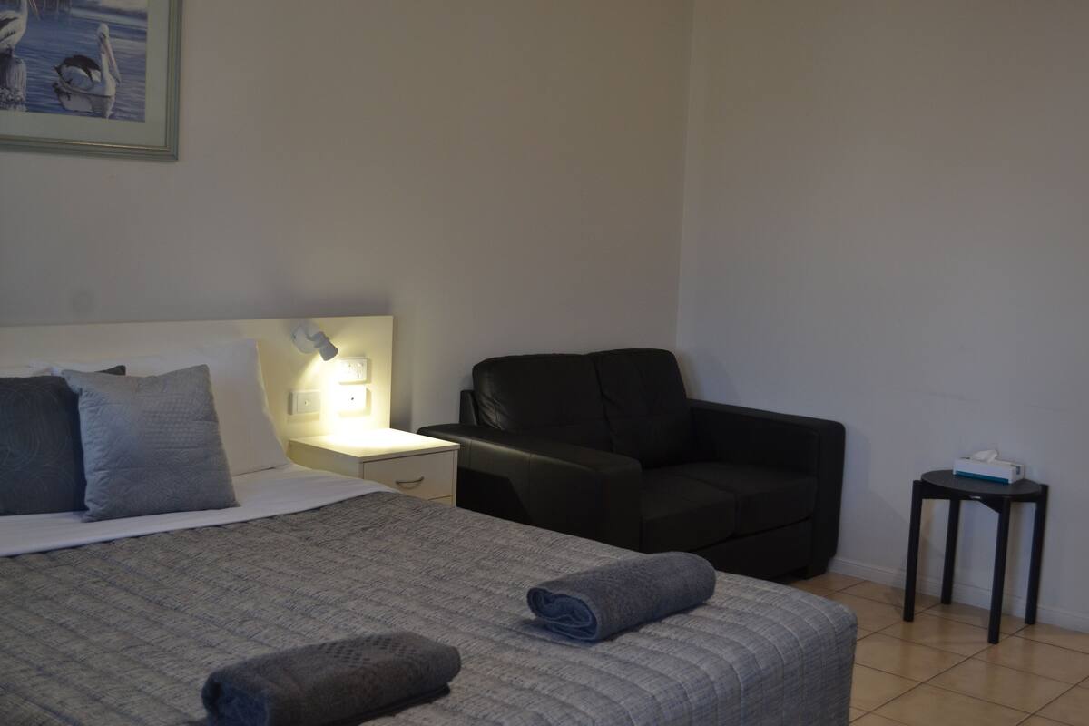 Et hotelværelse med en seng, et natbord med en lampe, en sort læderstol og et lille bord med en tissueske.