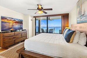 Un dormitorio con una cama grande, un ventilador de techo, un televisor sobre un aparador de madera y vista al océano a través de amplias ventanas.