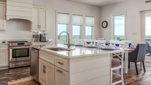 A modern kitchen with white cabinetry, stainless steel appliances, a large island with seating, and a view to the ocean. - Coastal Charm | Nightly stays starting 9/20! Pool (Dauphin Island)