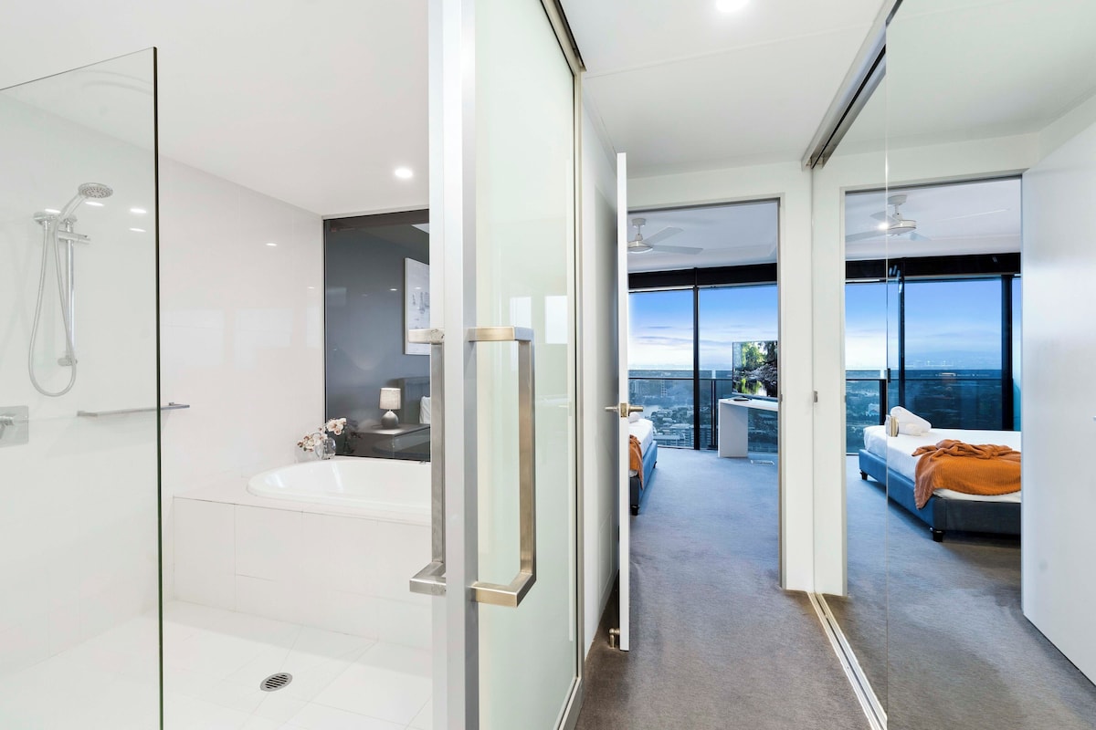 Ein modernes Hotelzimmer mit Duschkabine aus Glas, einer großzügigen Badewanne, einem Balkon mit Stadtblick und einem Bett mit Blick.