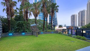 A well-maintained grassy area with palm trees, a stone structure, and modern buildings in the background. - Converted 2 Bedroom Apartments (No Parking) - WOWSTAY (Surfers Paradise)