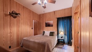 A bedroom with wooden paneling, a bed with a beige cover, a ceiling fan, a nightstand with a lamp, and a door.