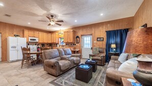 A spacious living area with a sofa, coffee table, and a dining table with chairs. A kitchen with wooden cabinets and a microwave is visible in the background.