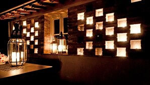A dimly lit room with a brick wall featuring illuminated square cutouts, a wooden ceiling, and a table with a candle and two lanterns.