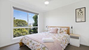 Un dormitorio con un ventanal grande, una cama con ropa de cama floral, una mesita de noche y un cartel optométrico enmarcado en la pared.