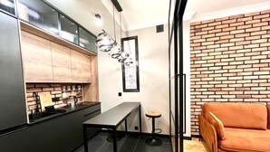 Una cocina moderna con gabinetes negros, encimera negra y una lámpara colgante. Hay un taburete pequeño y un sofá en la zona de estar contigua, con una pared de ladrillo.