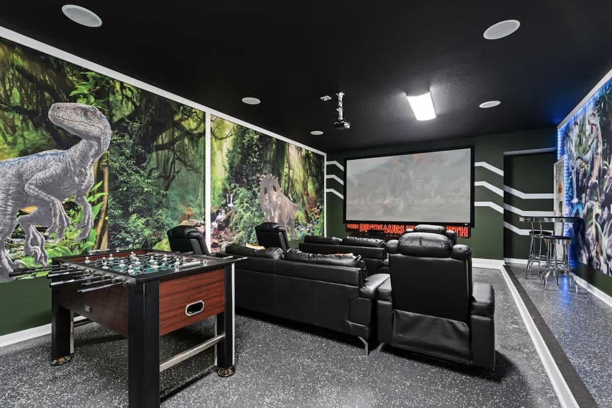 Een kamer met een tafelvoetbaltafel, een groot scherm, een bank en een muurschildering van dinosaurussen.