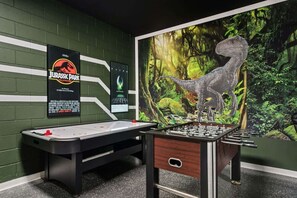 A room with a foosball table, a poster of a dinosaur, and a green brick wall.